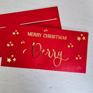 Könnte beinhalten: Rote Weihnachtskarte mit goldener Schrift "Merry Christmas Derry" und goldenen Sternen.