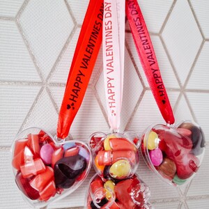 K&ouml;nnte beinhalten: Drei transparente Plastik-Valentinstags-Anh&auml;nger gef&uuml;llt mit roten und braunen S&uuml;&szlig;igkeiten. Die Anh&auml;nger sind in Form eines Herzens und eines B&auml;ren. Jeder Anh&auml;nger ist an einem roten, rosa oder wei&szlig;en Band befestigt, auf dem der Text "Froher Valentinstag" steht.
