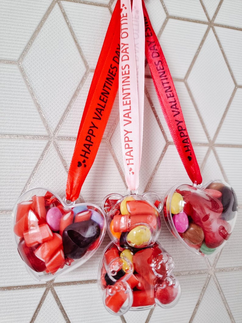 K&ouml;nnte beinhalten: Drei transparente Plastik-Valentinstags-Anh&auml;nger gef&uuml;llt mit roten und braunen S&uuml;&szlig;igkeiten. Die Anh&auml;nger sind in Form eines Herzens und eines B&auml;ren. Jeder Anh&auml;nger ist an einem roten, rosa oder wei&szlig;en Band befestigt, auf dem der Text "Froher Valentinstag" steht.