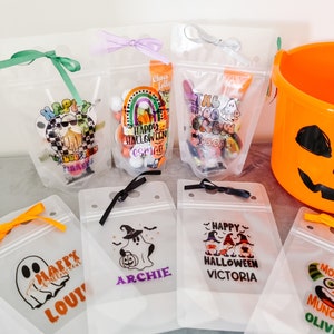 Könnte beinhalten: Sieben transparente Plastiktüten mit Reißverschluss, jede mit einem anderen Halloween-Motiv und einem personalisierten Namen verziert. Die Tüten sind mit Süßigkeiten und anderen Leckereien gefüllt.
