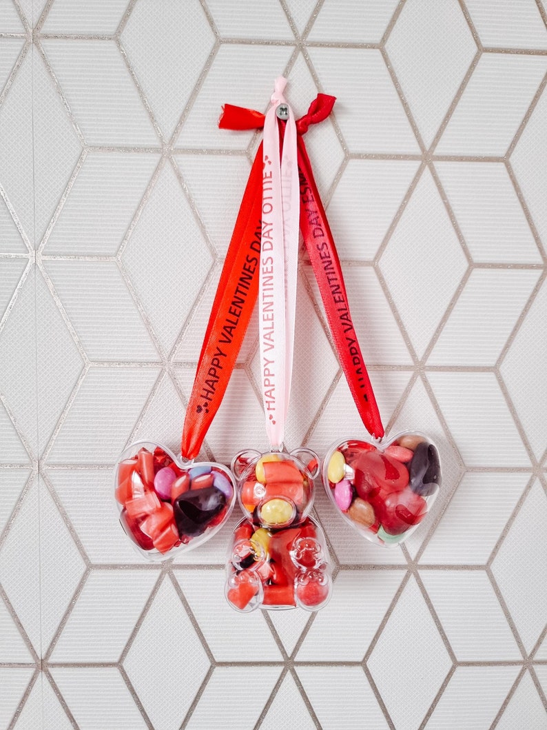 K&ouml;nnte beinhalten: Drei transparente Plastik-Valentinstags-Anh&auml;nger gef&uuml;llt mit bunten S&uuml;&szlig;igkeiten. Die Anh&auml;nger sind in Form eines Herzens, eines B&auml;ren und eines weiteren Herzens. Sie h&auml;ngen an roten und rosa B&auml;ndern mit dem Aufdruck "Happy Valentine's Day".