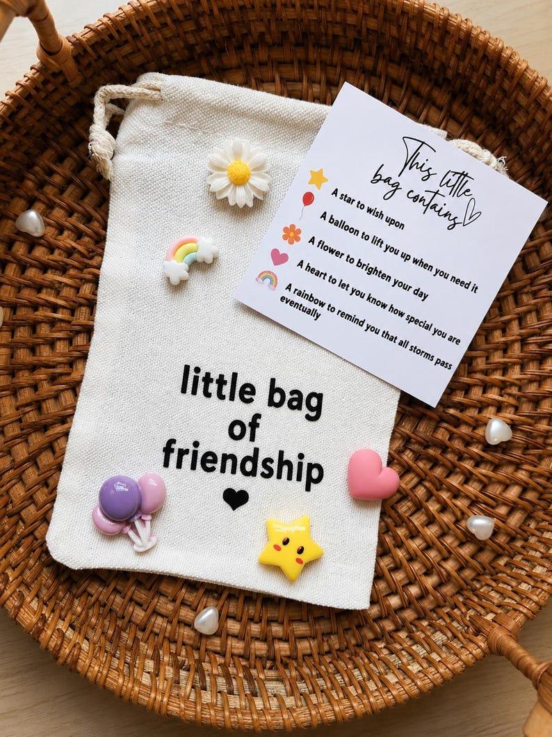 May include: A cream-colored drawstring pouch with the text 'little bag of friendship' and a small black heart. Attached are miniature charms: a white daisy, a rainbow, purple balloons, a pink heart, and a yellow star, plus a card listing the items.