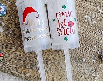May include: Two clear plastic snow poppers with white contents and long, clear handles. One has a Santa hat graphic and text that reads "Aera's Christmas Eve Snow Popper". The other says "Esmae Let it Snow" with green snowflake accents.