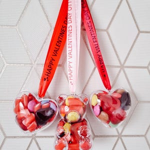 K&ouml;nnte beinhalten: Drei transparente Plastikbeh&auml;lter gef&uuml;llt mit bunten S&uuml;&szlig;igkeiten, in Form eines Herzens, eines B&auml;ren und eines weiteren Herzens. Jeder Beh&auml;lter h&auml;ngt an einem roten, rosa und wei&szlig;en Band mit dem aufgedruckten Text "Happy Valentines Day Ollie".