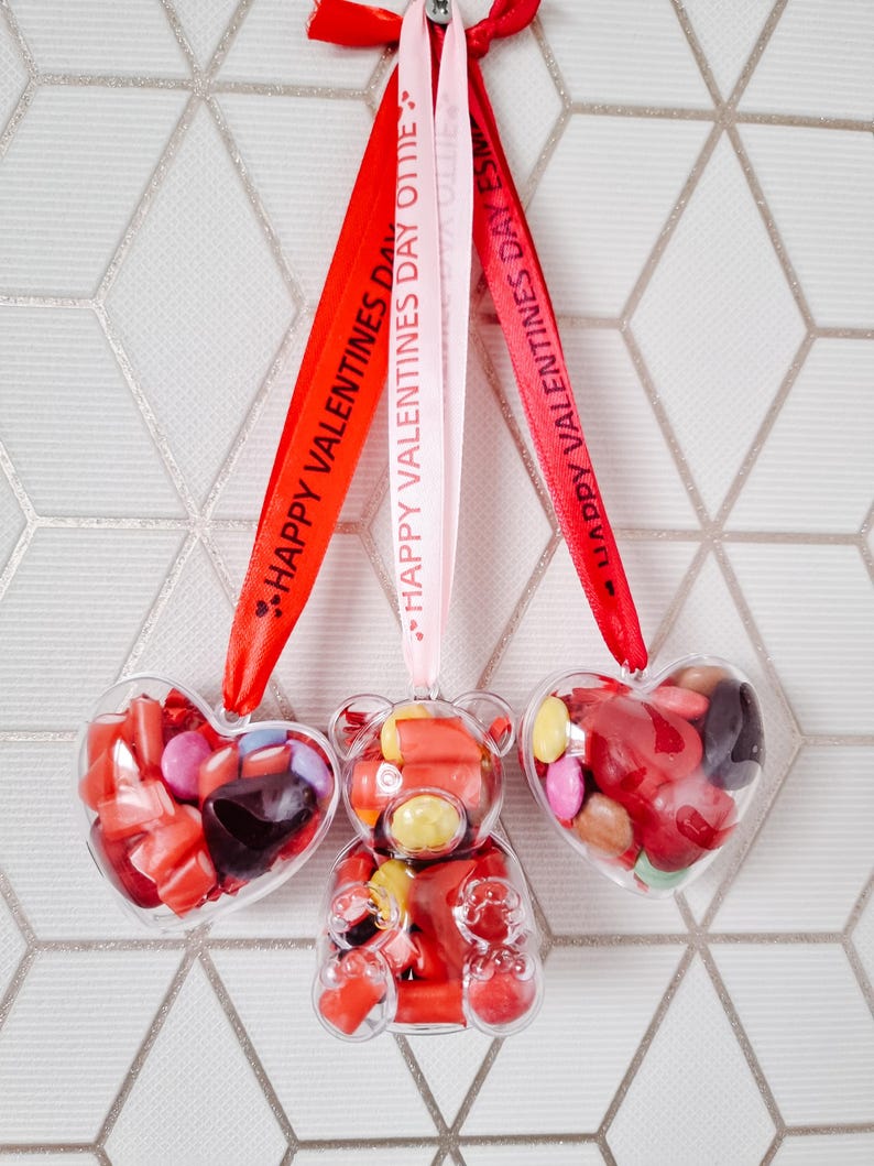 K&ouml;nnte beinhalten: Drei transparente Plastikbeh&auml;lter gef&uuml;llt mit bunten S&uuml;&szlig;igkeiten, in Form eines Herzens, eines B&auml;ren und eines weiteren Herzens. Jeder Beh&auml;lter h&auml;ngt an einem roten, rosa und wei&szlig;en Band mit dem aufgedruckten Text "Happy Valentines Day Ollie".