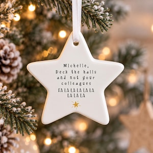 May include: A white star-shaped ceramic ornament hanging from a white ribbon. The text reads: Michelle, Deck the halls and not your colleagues, FA LA LA LA LA LA LA LA LA. A small gold star icon is printed below the text on the smooth surface.