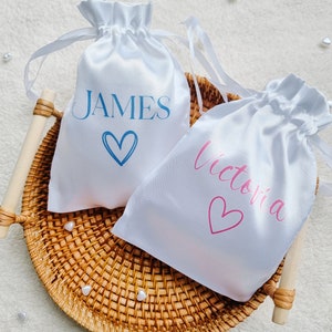 Könnte beinhalten: Zwei weiße Satin-Zugbeutel mit personalisierten Namen. Der Beutel links trägt den Namen "JAMES" in blau mit einem blauen Herzen. Der Beutel rechts trägt den Namen "Victoria" in rosa mit einem rosa Herzen.
