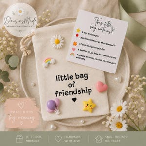 May include: A cream-colored fabric drawstring pouch with the text "little bag of friendship" printed in black. The pouch is decorated with small resin charms including a daisy, a rainbow, two balloons, and a yellow star, plus a card.