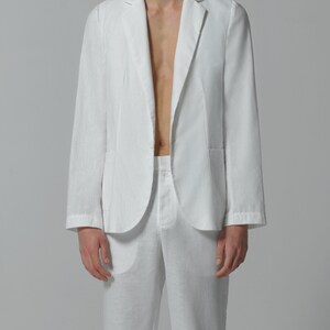 May include: A white linen blazer with a single button closure and a pocket on the left side. The blazer is unbuttoned and the person is wearing white linen pants.
