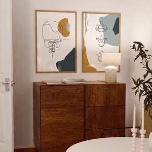 May include: Two framed abstract line art prints with a wooden dresser with three drawers, a lamp with a face design, and two candlesticks with pink candles. The prints feature a yellow, blue, and white color scheme.