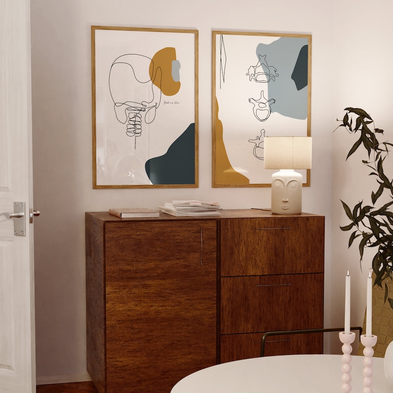 May include: Two framed abstract line art prints with a wooden dresser with three drawers, a lamp with a face design, and two candlesticks with pink candles. The prints feature a yellow, blue, and white color scheme.