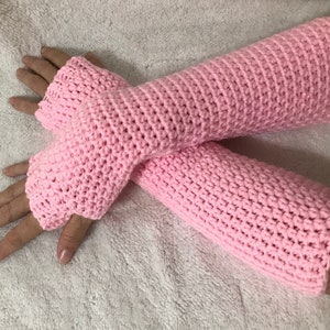 May include: A pair of light pink, crocheted arm warmers. The fingerless gloves are a solid color and have a textured stitch pattern. The arm warmers are displayed on a white surface.