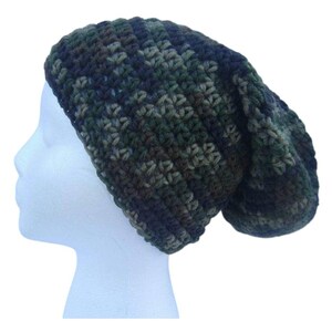May include: A camouflage-patterned crochet beanie hat in shades of green, brown, and navy blue. The hat is shown on a white mannequin head, showcasing its slouchy fit and textured stitch.
