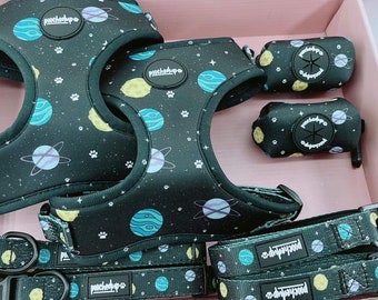 May include: A set of two black dog harnesses with a space-themed print, two matching leashes, and two poop bag holders. The harnesses and leashes feature a blue, yellow, and white planet design with a black background. The poop bag holders have a black background with a white star and planet design. The text "pouchd'up" is printed on the harnesses and leashes.
