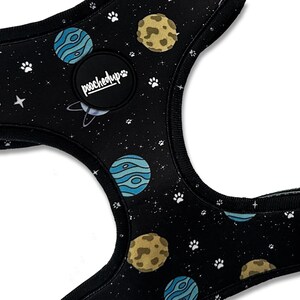May include: Black dog harness with a space-themed print featuring planets, stars, and paw prints. The harness has a black adjustable strap and a black buckle. The harness has a black patch with the text "pooched up" in white.