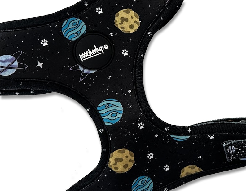 May include: Black dog harness with a space-themed print featuring planets, stars, and paw prints. The harness has a black adjustable strap and a black buckle. The harness has a black patch with the text "pooched up" in white.