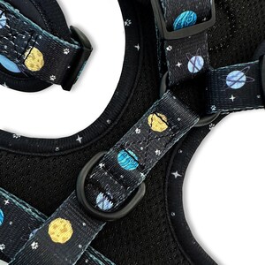 May include: A black dog harness with a blue and yellow space-themed pattern. The harness has a black adjustable buckle and a black D-ring.