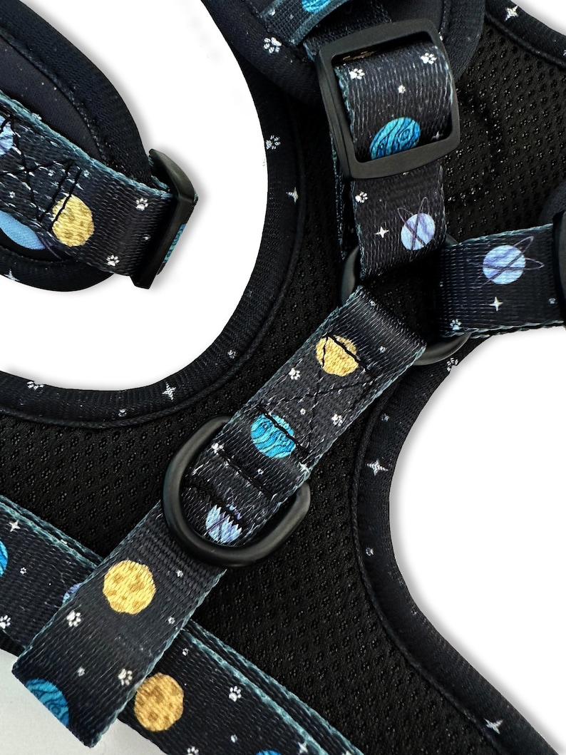 May include: A black dog harness with a blue and yellow space-themed pattern. The harness has a black adjustable buckle and a black D-ring.