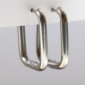 May include: A pair of silver-colored, square hoop earrings. The earrings are made of a shiny, metallic material and have a simple, modern design. The hoops are attached to a white surface.
