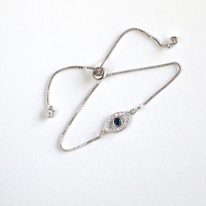 May include: A silver adjustable bracelet with a blue evil eye charm. The charm is surrounded by clear rhinestones.