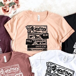 May include: Five t-shirts in different colours with a black and white graphic print of a sign that says "Scuttle's Thrift Shop" with a cartoon angel holding a shopping bag. The sign has a variety of items listed with prices, including "Dinglehopper 25p", "Sharfblatt £1.99", and "Set of 22 Thingamabobs £1.99".