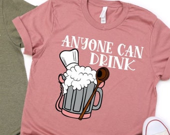 May include: Two t-shirts, one olive green and one dusty rose, each with the phrase "ANYONE CAN DRINK" in white script. A cartoon beer mug with a chef's hat and wooden spoon is printed below the text. White sneakers are visible.