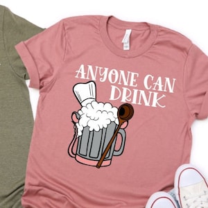 May include: Two t-shirts, one olive green and one dusty rose, each with the phrase "ANYONE CAN DRINK" in white script. A cartoon beer mug with a chef's hat and wooden spoon is printed below the text. White trainers are visible.
