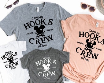 May include: A collection of gray, white, and peach-colored t-shirts and a onesie, all featuring the text "Captain Hooks Neverland Crew EST. 1802" with a Mickey Mouse-inspired skull and crossbones design. Sunglasses, a hat, and passports are also visible.