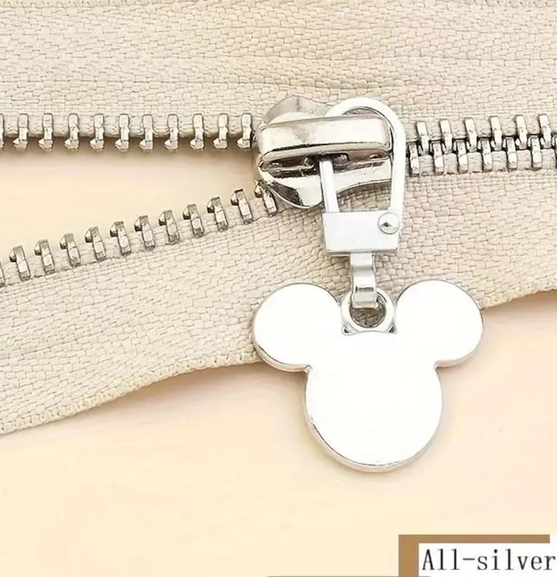 Op de afbeelding: Zilveren rits-trekker met een Mickey Mouse-kop ontwerp. De trekker is bevestigd aan een zilveren rits.
