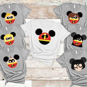May include: Several grey and white t-shirts with cartoon character designs. Each shirt displays a different character face, with black Mickey Mouse ears and a red, yellow, and black mask design. The shirts are arranged on a light wood surface.