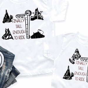 May include: White t-shirts featuring a black and red graphic that reads "Finally Tall Enough to Ride" with Disney-themed illustrations. The image also shows a pair of blue trainers and denim jeans.
