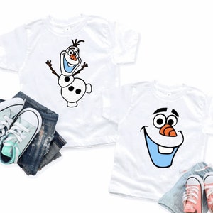 May include: Two white t-shirts featuring Olaf from Frozen. One shirt shows Olaf with arms raised, the other shows Olaf's face. The shirts are paired with denim shorts and sneakers in mint green and coral.