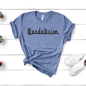 May include: A light blue t-shirt with the word "WandaVision" printed in black. The shirt is styled with a knot at the bottom and is paired with white sneakers. Small potted succulents are placed on a white wooden surface.