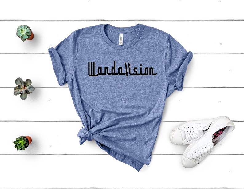 May include: A light blue t-shirt with the word "WandaVision" printed in black. The shirt is styled with a knot at the bottom and is paired with white sneakers. Small potted succulents are placed on a white wooden surface.