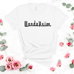 May include: White t-shirt with the word "WandaVision" printed in black. The shirt is surrounded by pink roses and green leaves on a white marble surface. The shirt is knotted at the bottom.