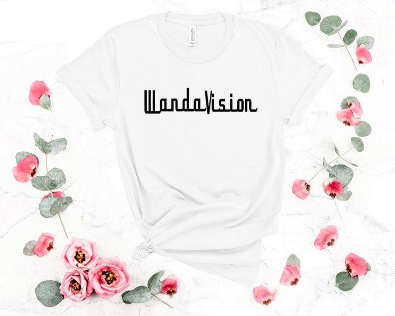 May include: White t-shirt with the word "WandaVision" printed in black. The shirt is surrounded by pink roses and green leaves on a white marble surface. The shirt is knotted at the bottom.
