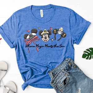 May include: A heather blue short-sleeve t-shirt featuring a graphic of Mickey Mouse wearing a captain's hat, a pair of nautical-themed mouse ears, and a large red bow. The text "Where Magic Meets the Sea" is printed in black cursive script.