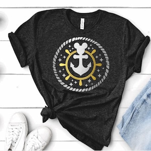 May include: A black t-shirt featuring a gold and white anchor design. The anchor is encircled by a rope circle and stars. The anchor has a Mickey Mouse head on top.