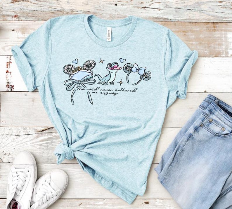 Może przedstawiać: Jasnoniebieski melanżowy t-shirt z nadrukiem graficznym przedstawiającym małą jaszczurkę, dwie pary ozdobnych uszu z kokardkami oraz napis "The cold never bothered me anyway" wykonany kursywą na środku klatki piersiowej.