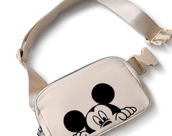 May include: Beige fanny pack featuring a black Mickey Mouse graphic. The bag has a zippered main compartment and an adjustable strap with a beige buckle. The strap and bag are made of a smooth, durable material.
