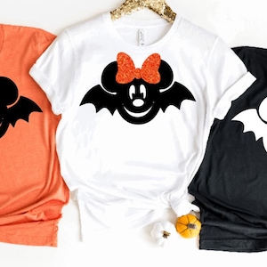 May include: Three t-shirts in orange, white, and black, each with a Halloween design. The design is a cartoon bat with mouse ears. The white shirt has a glittery orange bow. Pumpkins and decorative gourds are scattered around the shirts.