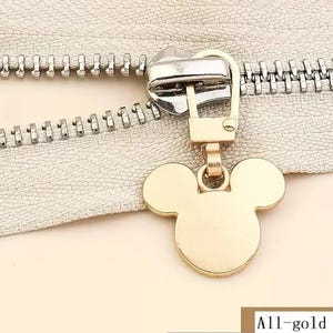 Op de afbeelding: Gouden rits-trekker met een Mickey Mouse-ontwerp. De rits-trekker is bevestigd aan een zilveren rits.