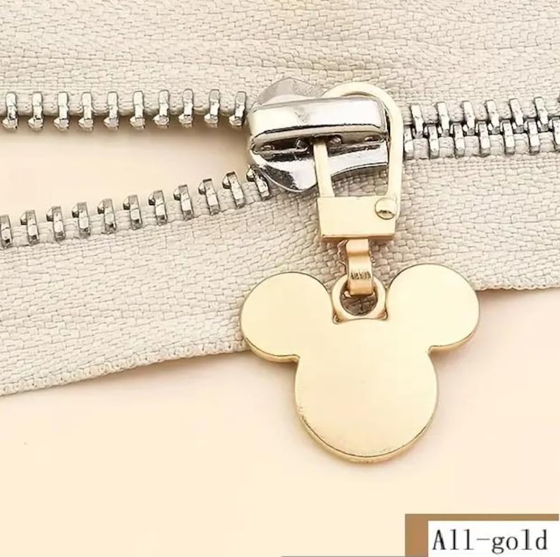 Op de afbeelding: Gouden rits-trekker met een Mickey Mouse-ontwerp. De rits-trekker is bevestigd aan een zilveren rits.