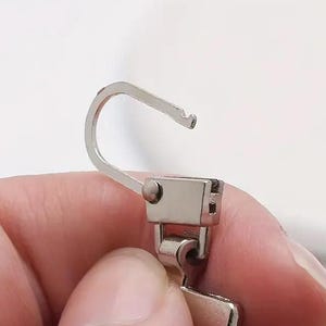 Op de afbeelding: Een zilveren rits met een haak en een schuif. De schuif heeft een klein metalen lipje aan de zijkant.