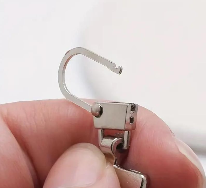 Op de afbeelding: Een zilveren rits met een haak en een schuif. De schuif heeft een klein metalen lipje aan de zijkant.