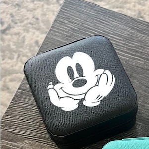 Puede incluir: Estuche de viaje cuadrado negro con un diseño blanco de Mickey Mouse. El estuche tiene una superficie texturizada y una cremallera negra. El diseño de Mickey Mouse está centrado y en una pose clásica.