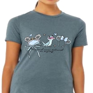 Może przedstawiać: Melirowany, łupkowoniebieski t-shirt z nadrukiem graficznym przedstawiającym jasnoniebieską jaszczurkę, dwie pary ozdobnych uszu ze śnieżynkami i kokardkami oraz kursywny napis "The cold never bothered me anyway" wyśrodkowany na klatce piersiowej.