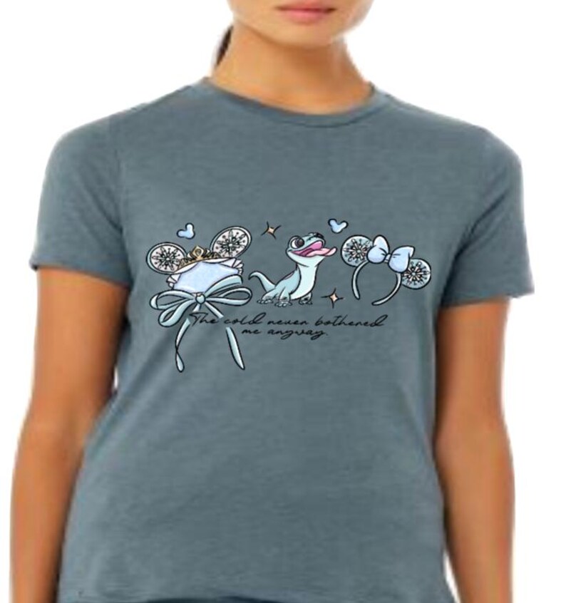 Może przedstawiać: Melirowany, łupkowoniebieski t-shirt z nadrukiem graficznym przedstawiającym jasnoniebieską jaszczurkę, dwie pary ozdobnych uszu ze śnieżynkami i kokardkami oraz kursywny napis "The cold never bothered me anyway" wyśrodkowany na klatce piersiowej.