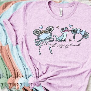 Może przedstawiać: Jasnofioletowy melanżowy t-shirt z grafiką przedstawiającą jaszczurkę, dwie pary mysich uszu oraz napis "The cold never bothered me anyway". Wz&oacute;r zawiera koronę, kokardy i małe iskierki w odcieniach niebieskiego, białego i złotego.