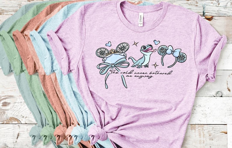 Może przedstawiać: Jasnofioletowy melanżowy t-shirt z grafiką przedstawiającą jaszczurkę, dwie pary mysich uszu oraz napis "The cold never bothered me anyway". Wz&oacute;r zawiera koronę, kokardy i małe iskierki w odcieniach niebieskiego, białego i złotego.